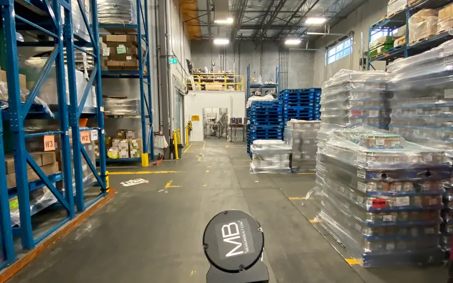 Warehouse aisle with pallet racking and stacked goods, with a MeasureBuilt laser scanner on the floor.